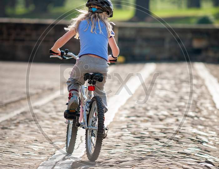 Young Girl Riding Bike Away From Camera Along A Cobbled Path