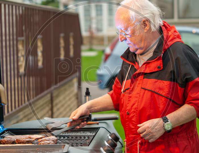Elderly Man In A Red Coat Cooking Meat On A Bbq