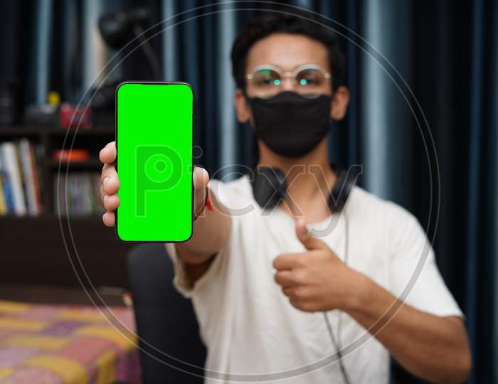 Young Indian Boy Wearing A Black Mask Holding A Phone With Green Screen, Showing Thumbs Up In The Background.