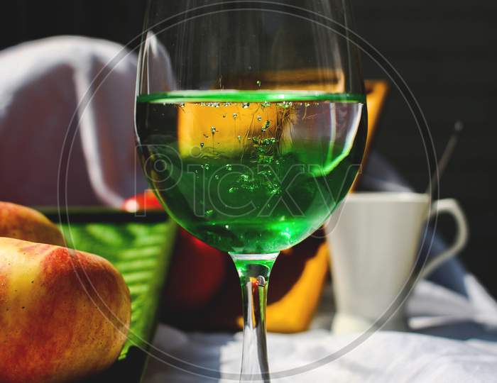 Pouring Squash Into A Glass Of Water, It Is Kept On A White Sheet With Fruits, Trays And A Ceramic Cup In The Background.