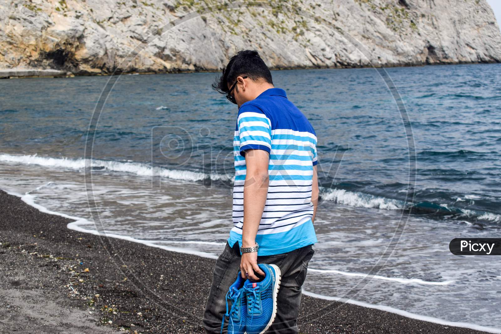Santorini / Greece: April 26, 2019: Man wearing blue T-Shirt and black Jeans walking on black sand beach with shoes in his hands