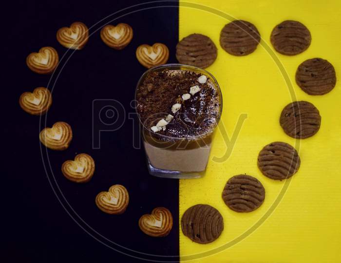 Dalgona coffee garnished with white chocolate and coffee powder on a black and yellow background decorated with biscuits.