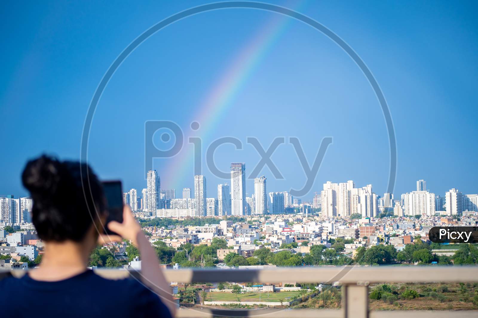 Young Indian Girl Wearing Hair In A Bun And With Blue Dress Photographing Rainbow Over Gurgaon Delhi Noida Cityscape On A Monsoon Day