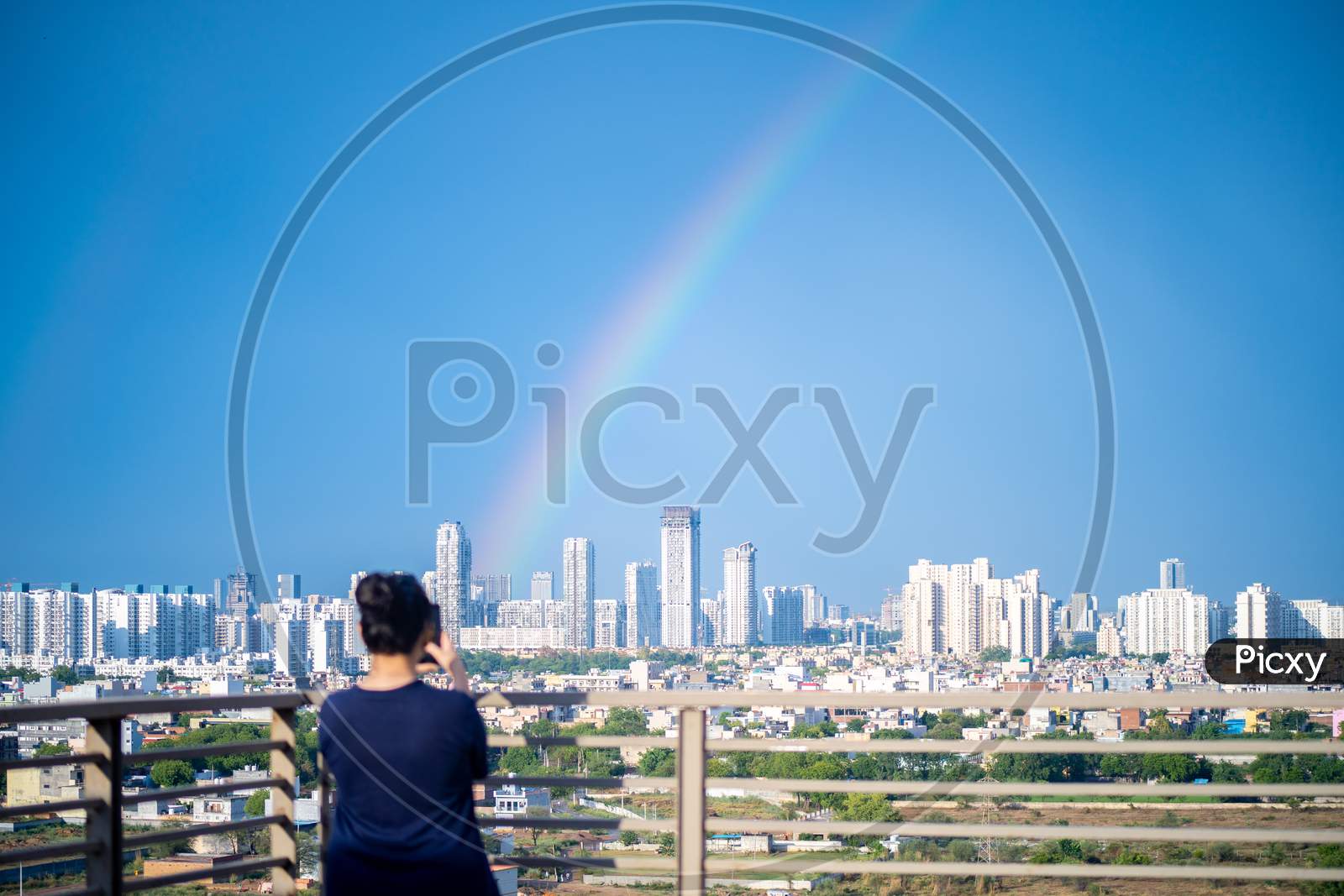Young Indian Girl Wearing Hair In A Bun And With Blue Dress Photographing Rainbow Over Gurgaon Delhi Noida Cityscape On A Monsoon Day