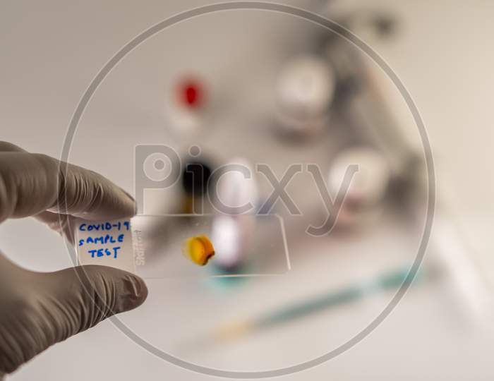 Photo concept shows Covid-19 sample analysis using a microscopic glass slide, with chemicals, syringe, and pipette in the blurred background.
