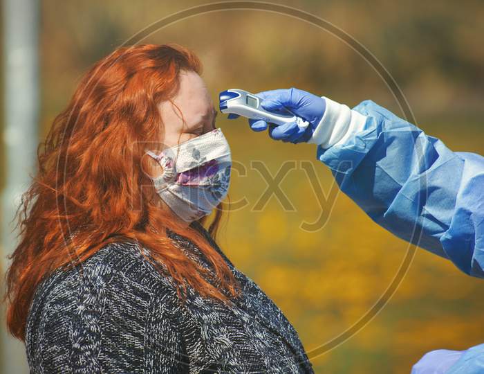 Close-up portrait of woman with cloth face mask being measured by a digital thermometer held in the medic's hand.