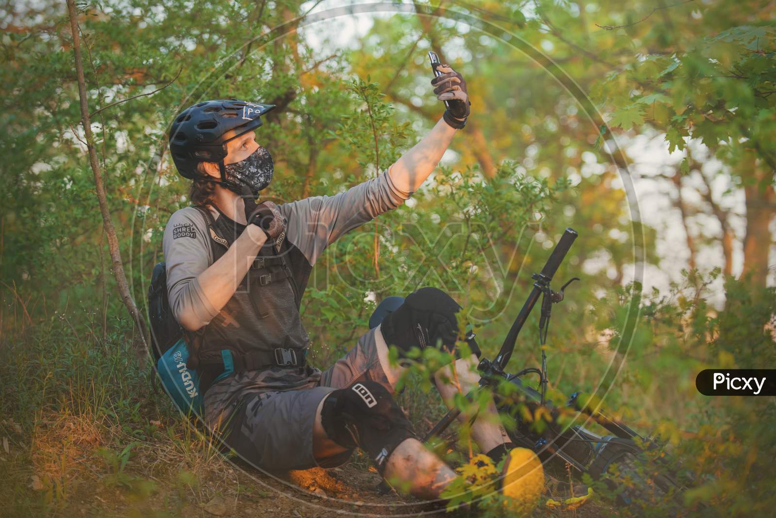 Cyclist wears a cloth face mask (due to mandatory wearing of face protection against the corona virus) with a bike lying next to him in the woods taking a selfie.