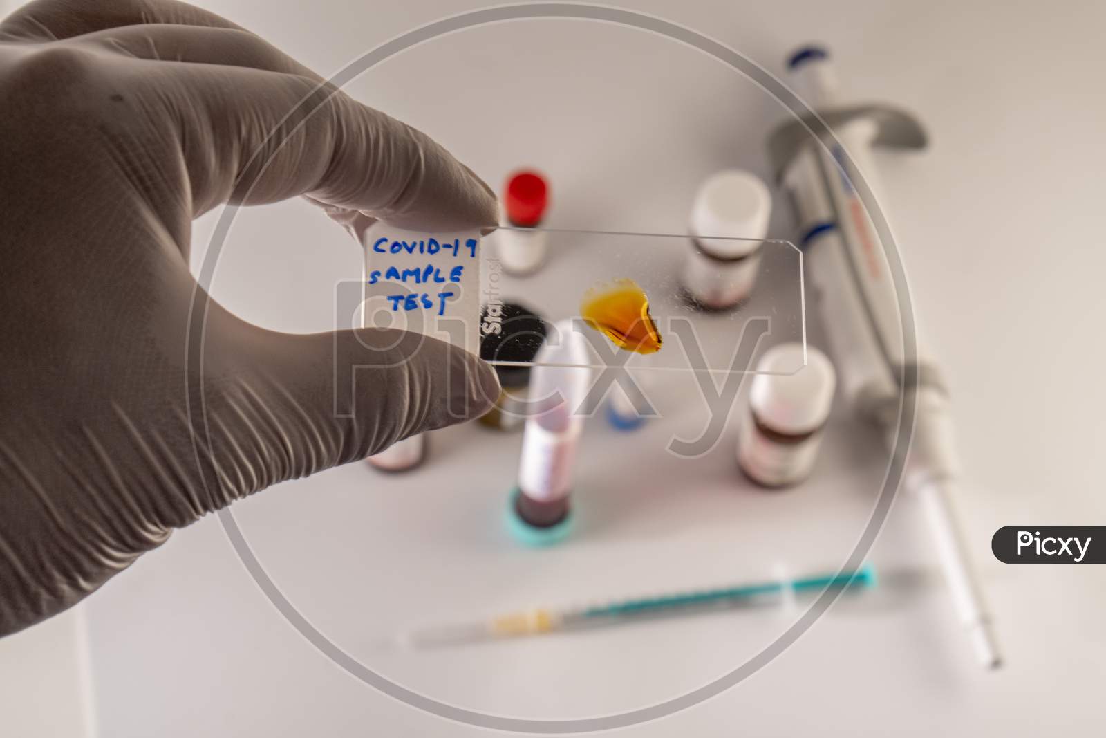 Photo concept shows Covid-19 sample analysis using a microscopic glass slide, with chemicals, syringe, and pipette in the blurred background.