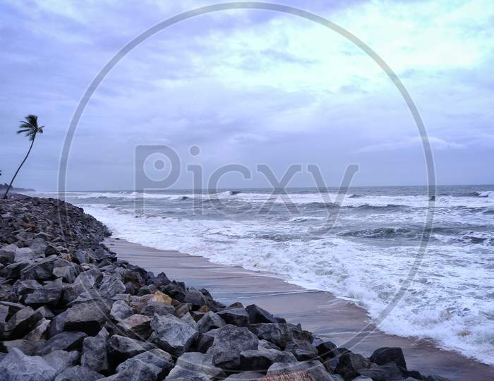 A Picture Of Beach With Rocks Along With A Coconut Tree On The Shore And Cloudy Sky On Background. The Sea Is Violent With Strong Waves In This Picture. Kappil Beach (Varkala Town), Kerala.