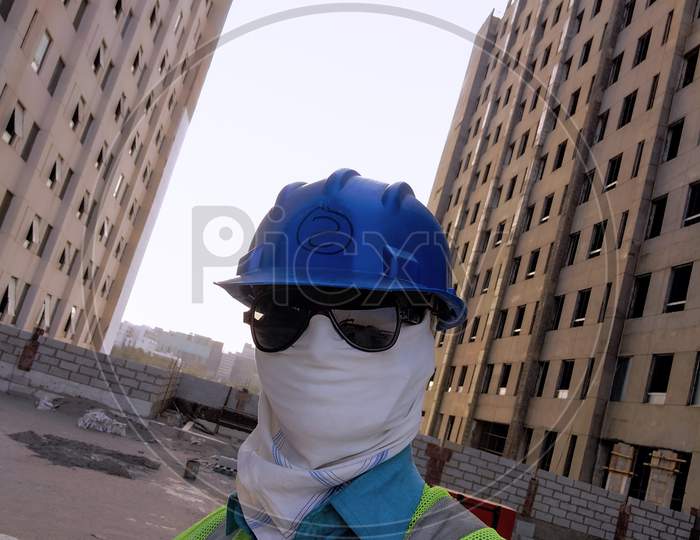 Construction Worker Covering Cloth On Face As Protective Mask Along With Other Safety Equipment, Under Construction Apartments In Background.