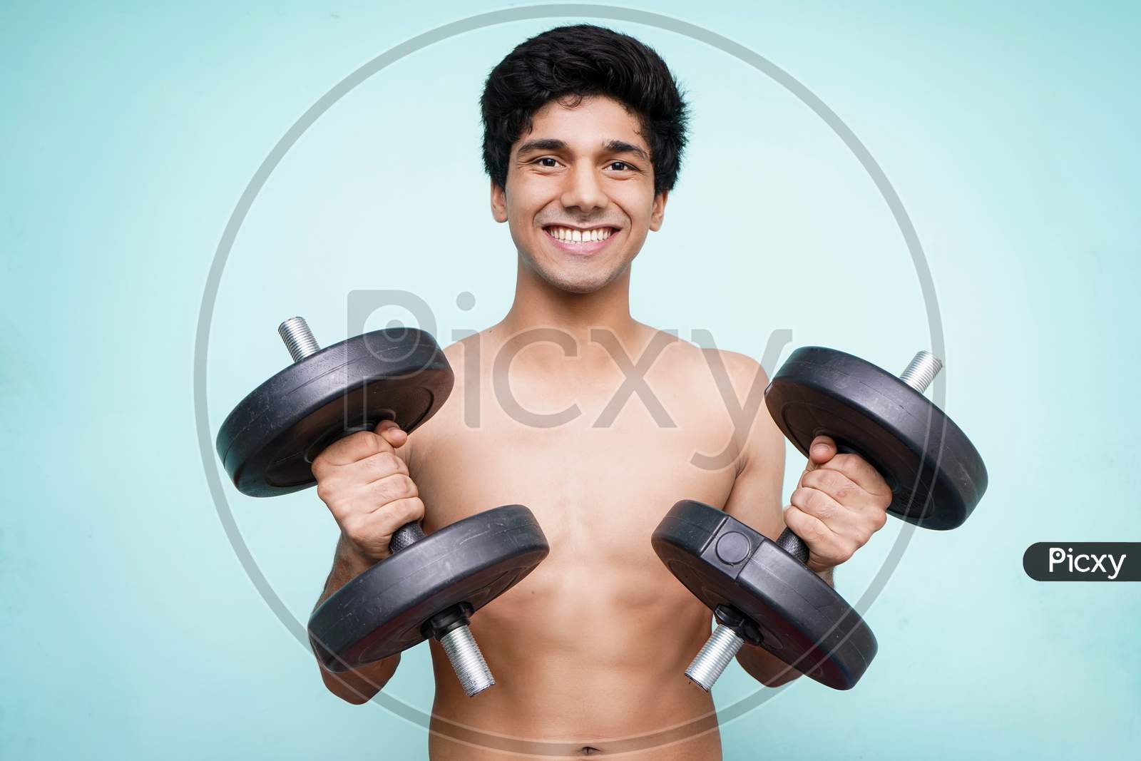 Young Handsome Asian Boy Holding Dumbbells On Both The Hands Looking Into The Camera While Smiling. Standing In Front Of A Blue Wall.