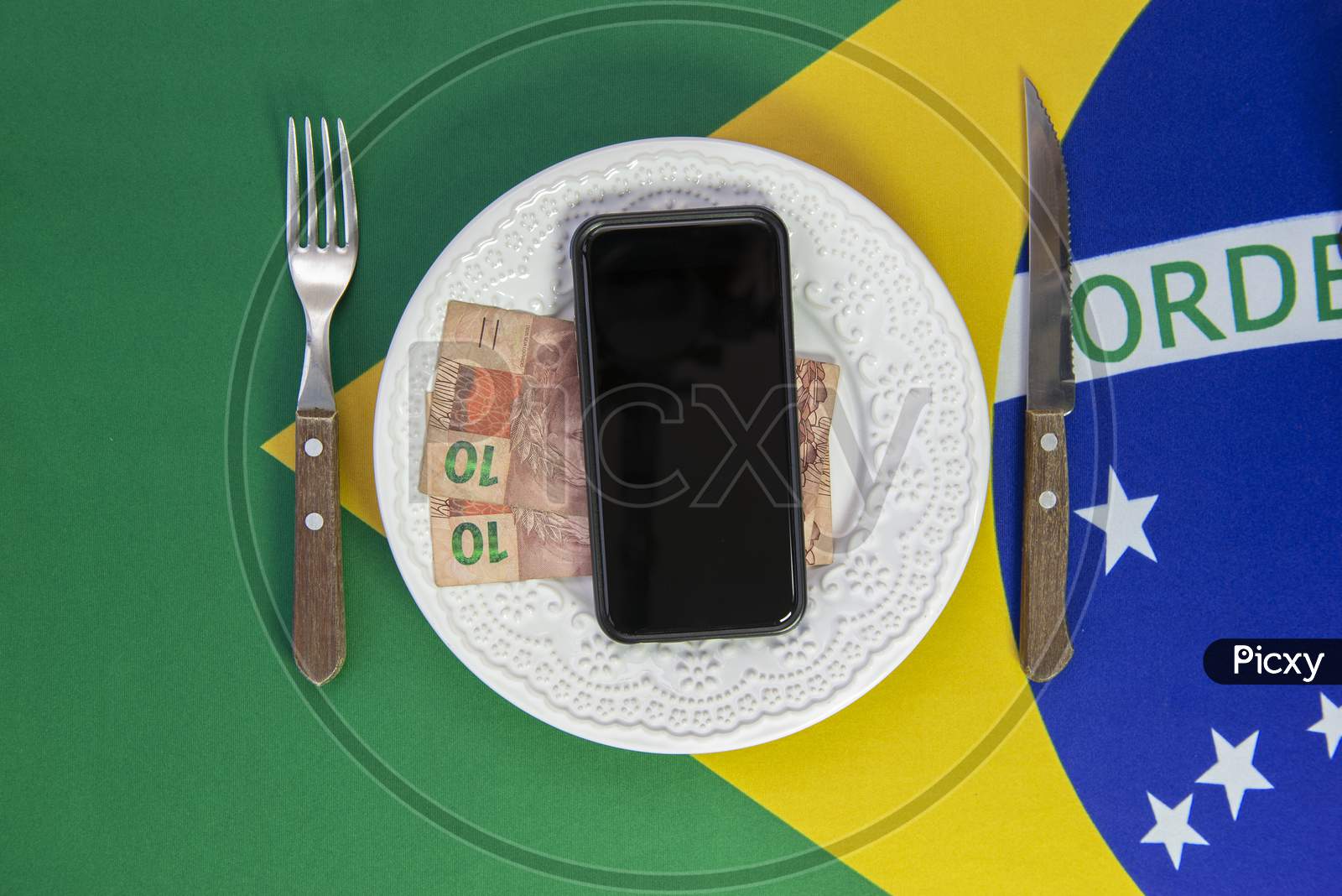 Top View Of Cellular Over Food Plate Next To Cutlery And Brazilian Flag In The Background. Mobile Phone With Black Screen.