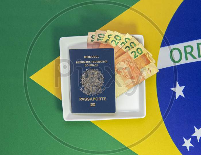 Brazilian Passport With Real Banknotes On A White Plate With The Flag Of Brazil In The Background. Cutlery Next To The Plate.