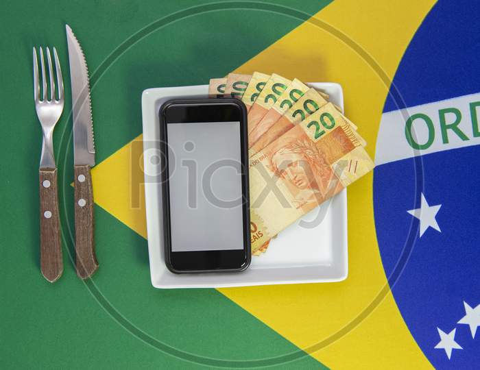 Top View Of Cellular Over Food Plate Next To Cutlery And Brazilian Flag In The Background. Mobile Phone With Blank Screen.