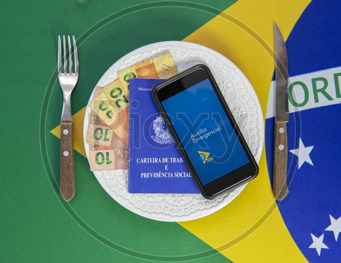 Money And Cell Phone With The Image Of The Emergency Aid Plan Brazilian Dish Of Food. Concept Of Social Aid To The Neediest. Background Brazilian Flag.