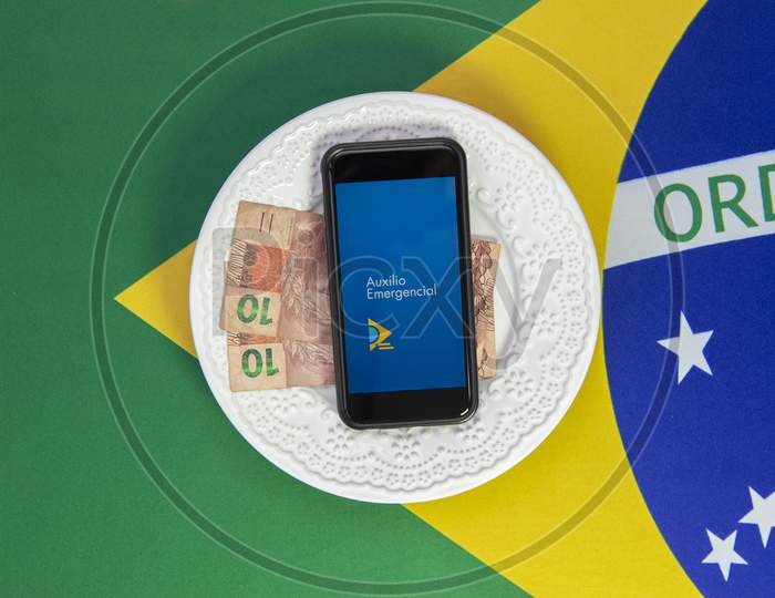 Money And Cell Phone With The Image Of The Emergency Aid Plan Brazilian Dish Of Food. Concept Of Social Aid To The Neediest. Background Brazilian Flag.
