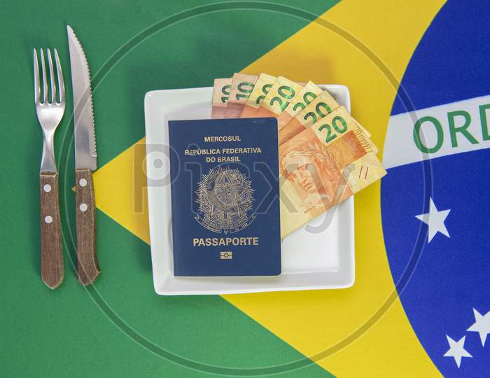 Brazilian Passport With Real Banknotes On A White Plate With The Flag Of Brazil In The Background. Cutlery Next To The Plate.