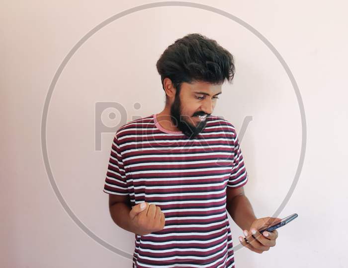 Indian Beard Man Looking At His Mobile Phone And Giving Reactions. Man Is Dressed In Stripped T-Shirt. Isolated On White Background.