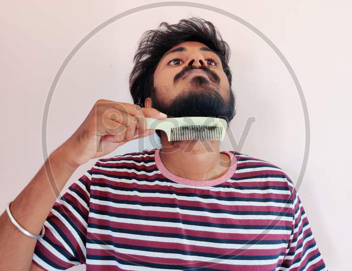 Young Indian Man Comb His Beard And Moustache On White Background. Man Is Dressed In Stripped T-Shirt. Daylight