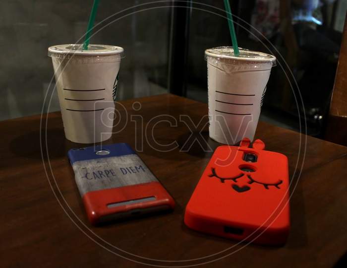 Two Smartphones With Back Covers Kept On A Brown Wooden Table Along With Two Disposal Coffee Cups.
