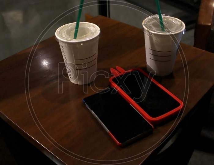 Two Smart Phones With Back Covers Kept On A Brown Wooden Table Along With Two Disposal Coffee Cups.