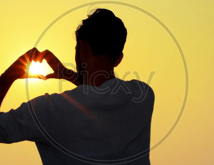 Silhouette Golden Rays Of Sun Through Hand Making Heart Shape Of A Young Man At Sunset .