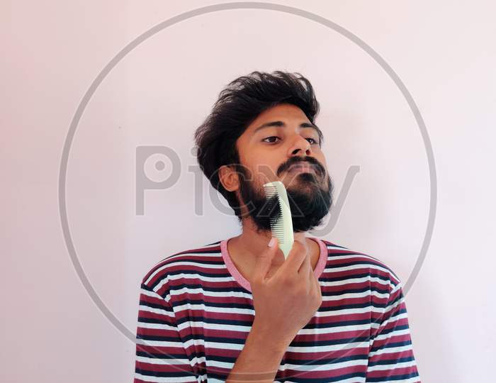 Young Indian Man Comb His Beard And Moustache On White Background. Man Is Dressed In Stripped T-Shirt. Daylight