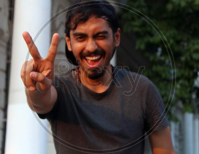 Young Man Winking And Smiling While Showing Peace Sign With His Fingers.
