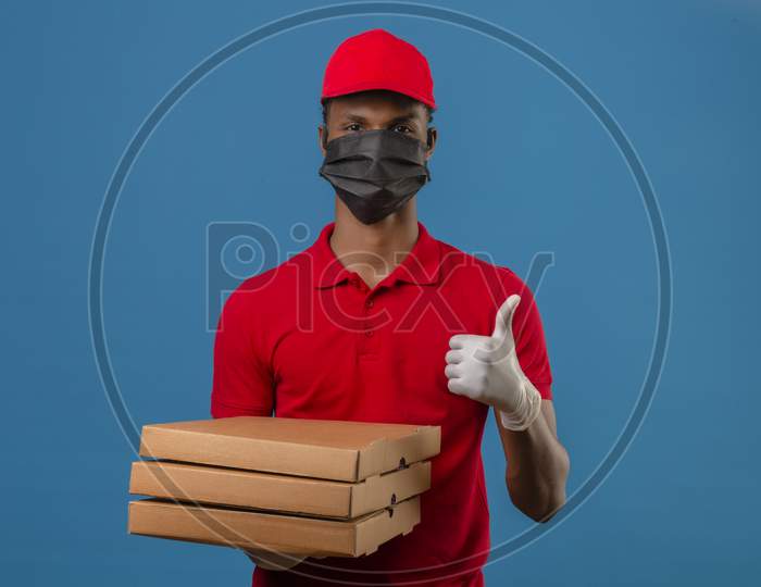 Young African American Delivery Man Wearing Red Polo Shirt And Cap In Protective Mask And Gloves Standing With Stack Of Pizza Boxes Showing Thumbs Up Over Isolated Blue Background