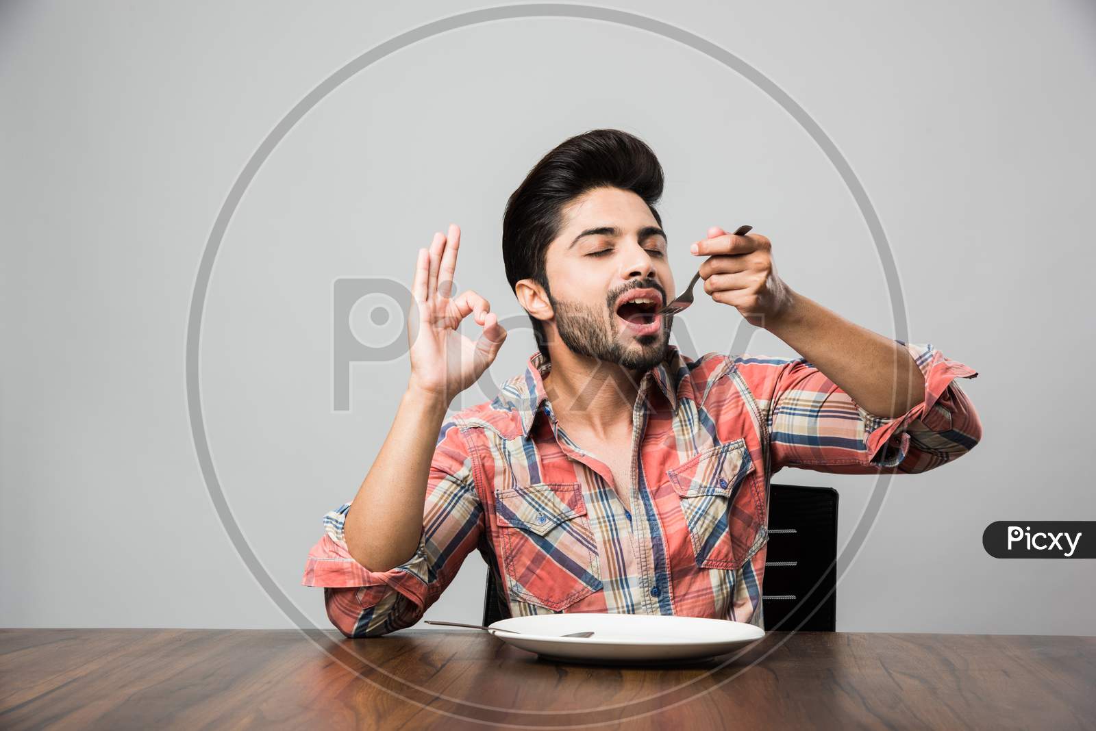 empty plate and Indian man with beard holding spoon and fork, wearing checkered shirt and sitting at table
