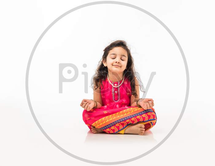 Portrait of cute little Indian girl model practicing and posing for pranayama or meditation while sitting isolated over white ba