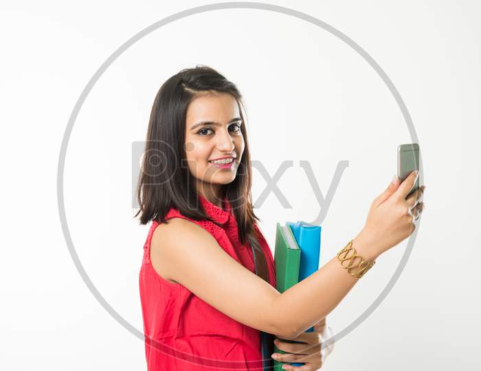 Pretty Indian/Asian College Girl holding books and clicking selfie with smartphone, standing isolated over white background