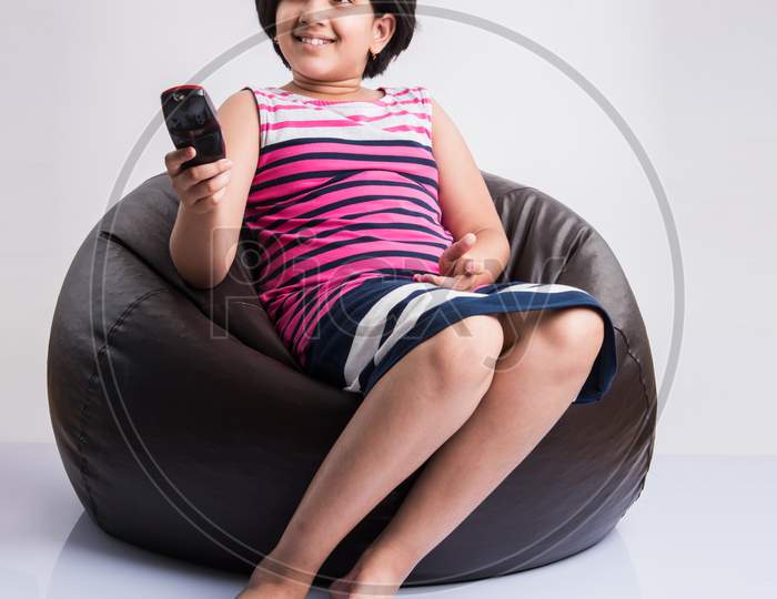Indian small girl watching Television / TV while holding remote control, sitting over bean bag