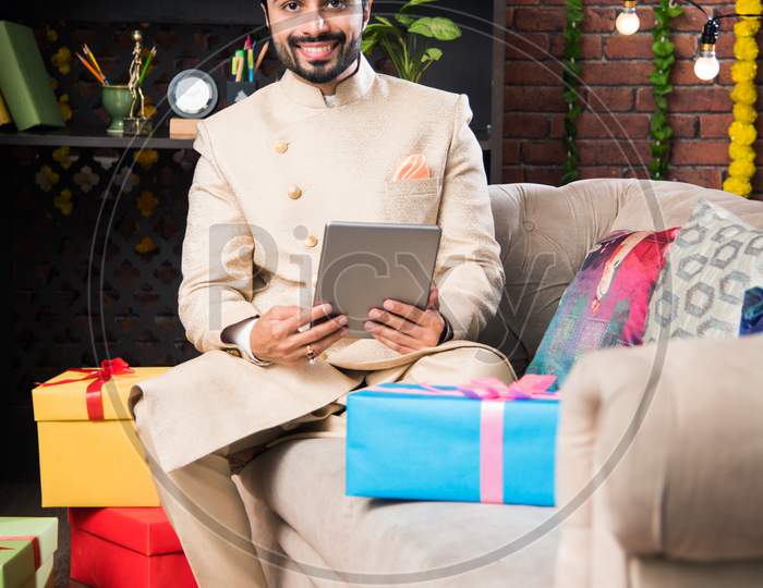 Indian man using tablet touchscreen computer on festival day while wearing traditional outfit. sitting on sofa with gifts and sw