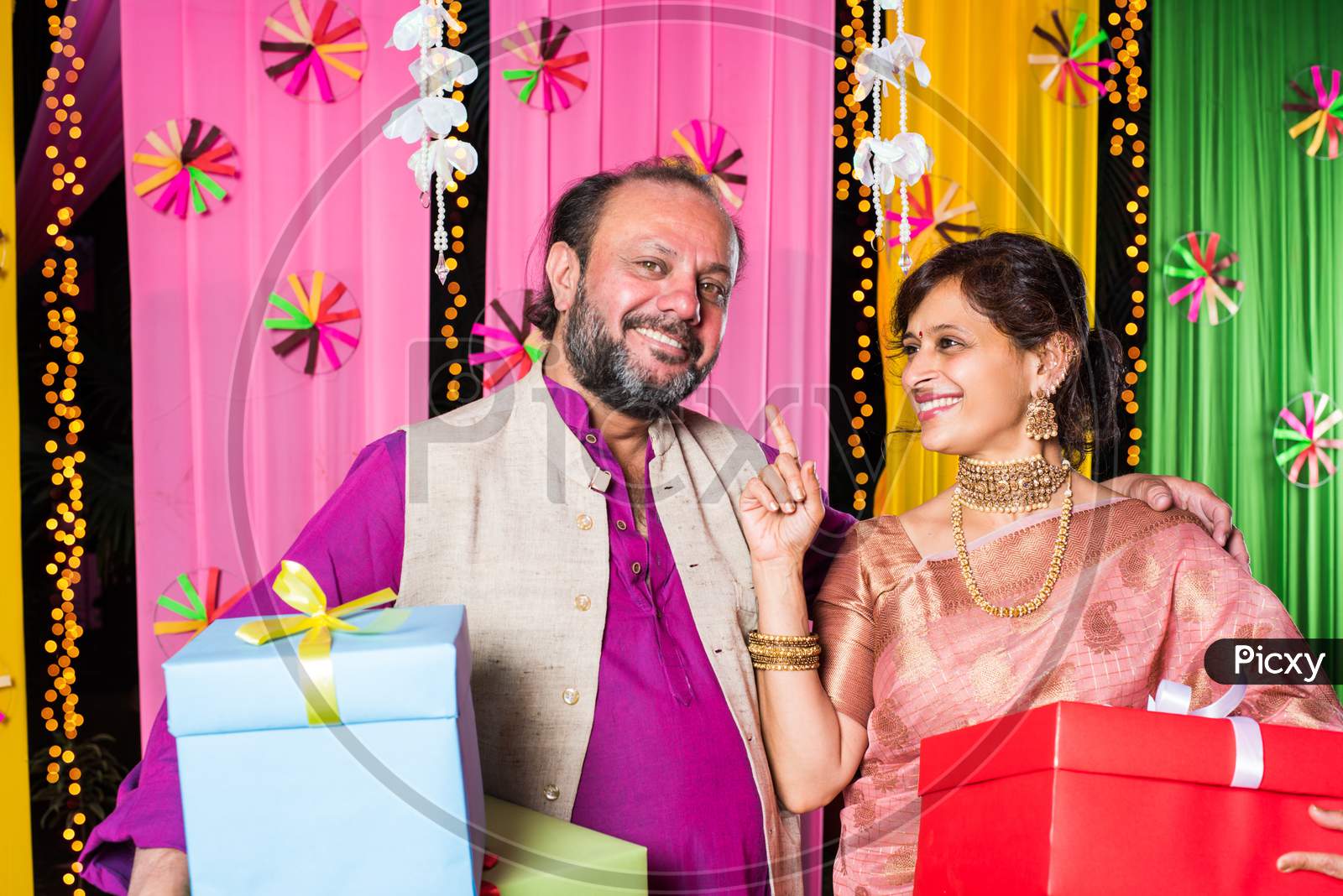 Mid age Indian couple holding Diwali Gift Boxes while standing against decorative background