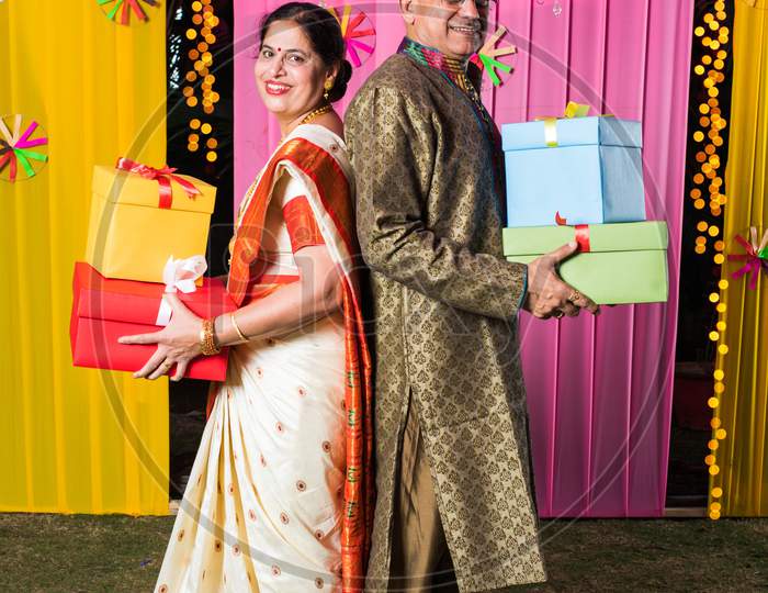 Senior Indian couple holding Diwali Gift Boxes while standing against decorative background