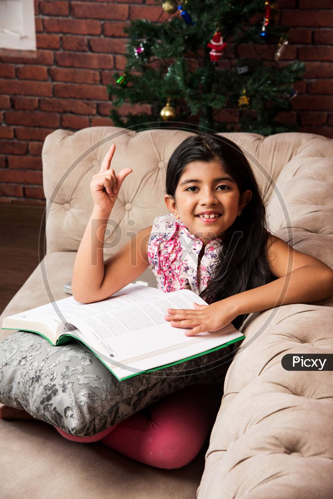 Cute little Indian/asian girl reading book while sitting sofa or couch at home