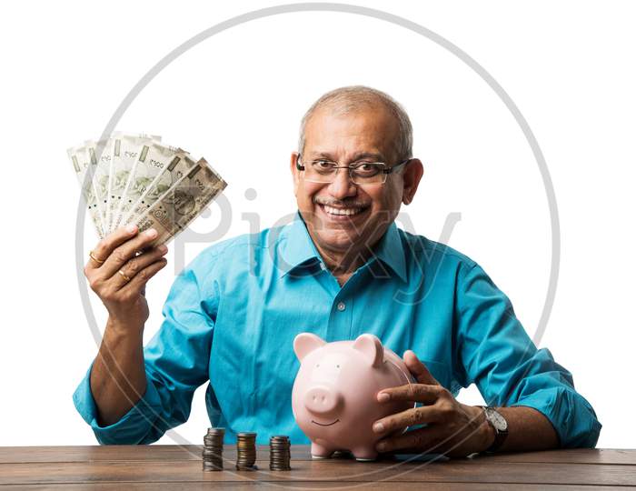 senior Indian/asian man and savings concept - with piggy bank, currency notes, 3d house model, money fan, pile of coins. sitting