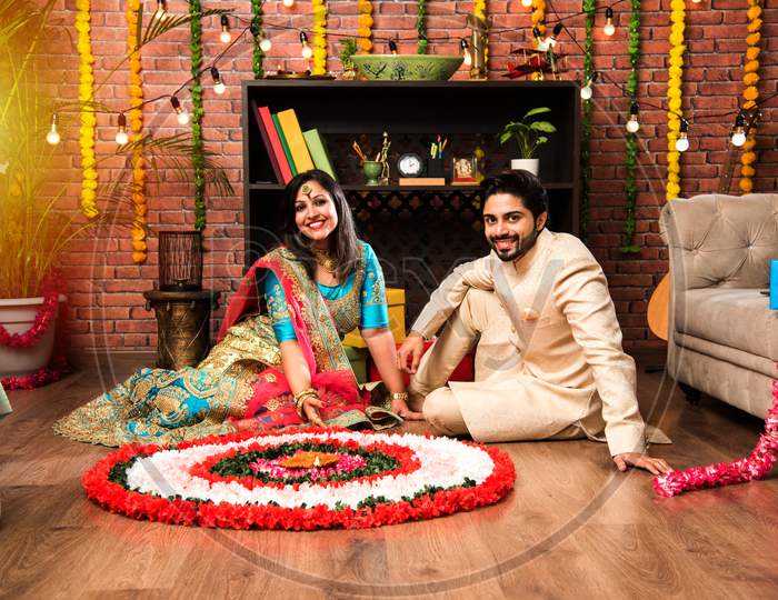 Indian couple making flower Rangoli on Diwali or Onam Festival, taking selfie or holding sweets
