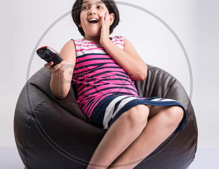Indian small girl watching Television / TV while holding remote control, sitting over bean bag