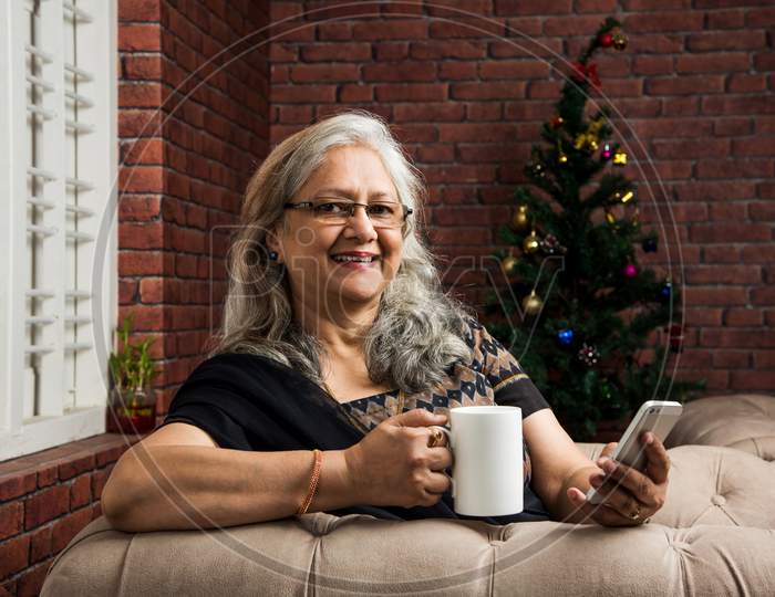 Old indian/asian lady or women using smartphone with a cup of coffee while sitting on sofa or couch at home
