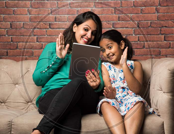 Indian mother and daughter on video call using using tablet computer, waving hands while sitting on sofa/couch