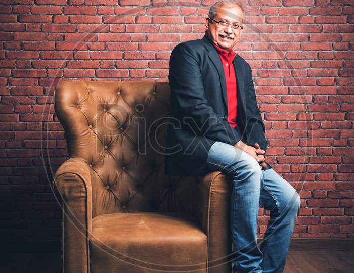 stylish senior indian/asian man in suit sitting on sofa against red brick background, moody lighting