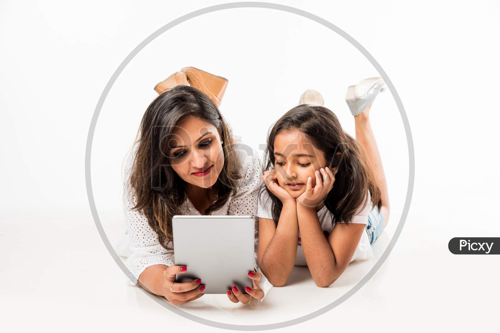 Indian mother and daughter lying on floor with book, laptop or tablet computer studying or readying story or playing games