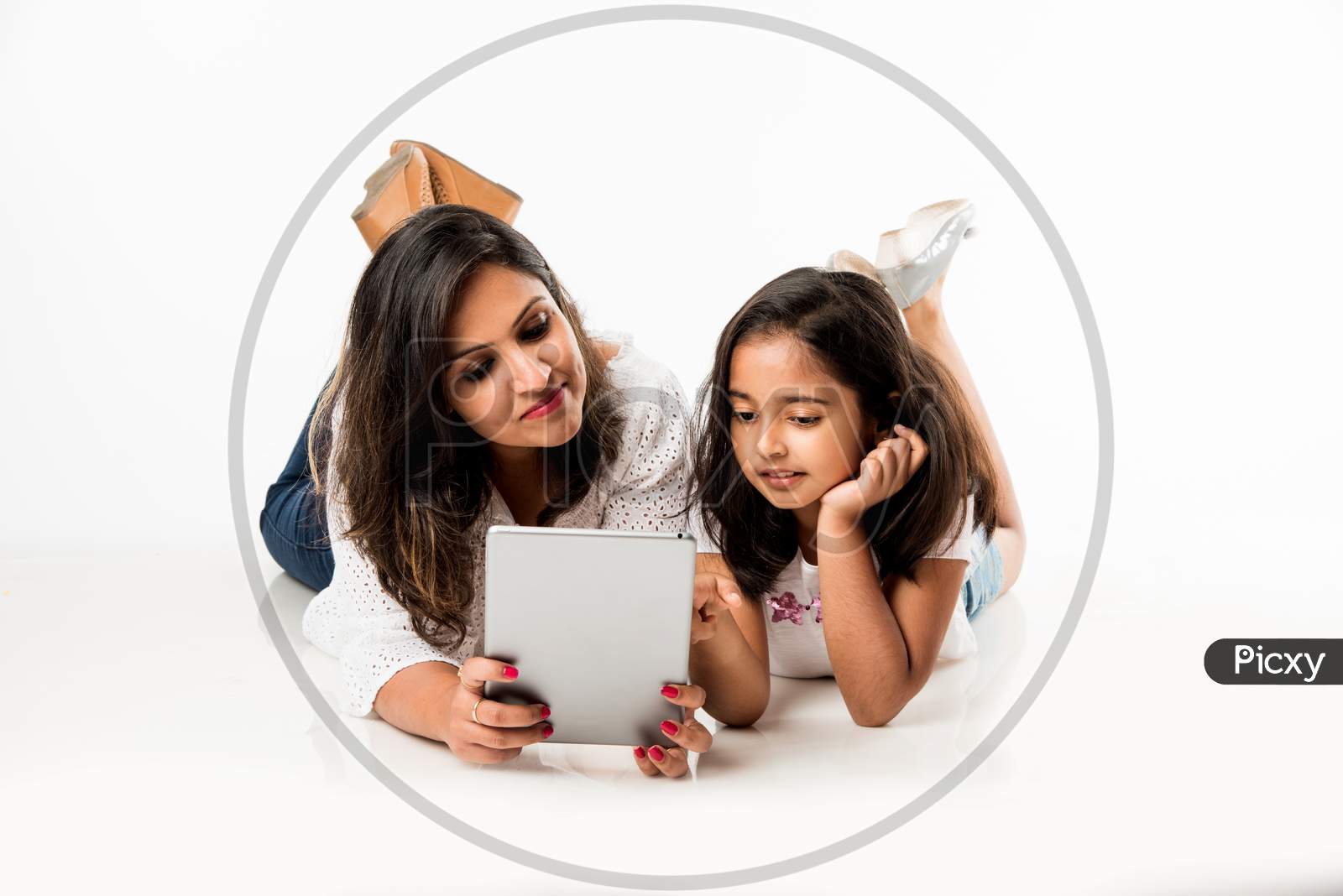 Indian mother and daughter lying on floor with book, laptop or tablet computer studying or readying story or playing games