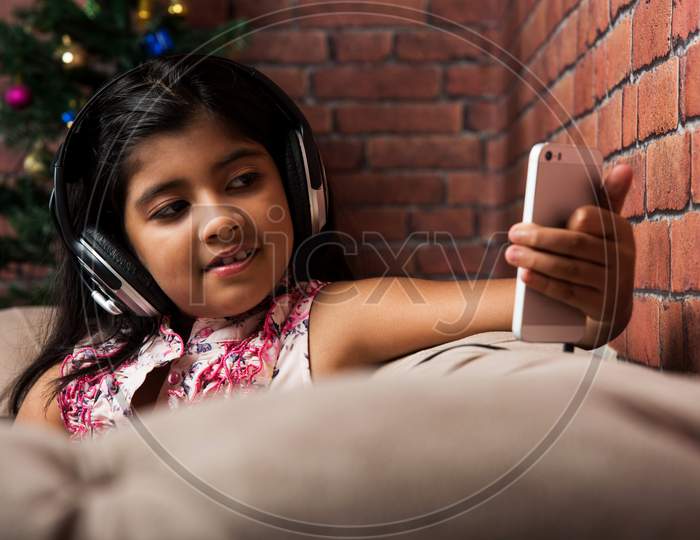 cute little indian/asian small girl listening to music using headphones or earphone on smartphone, sitting on couch or sofa at h