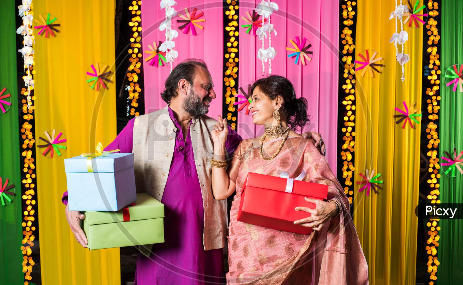 Mid age Indian couple holding Diwali Gift Boxes while standing against decorative background