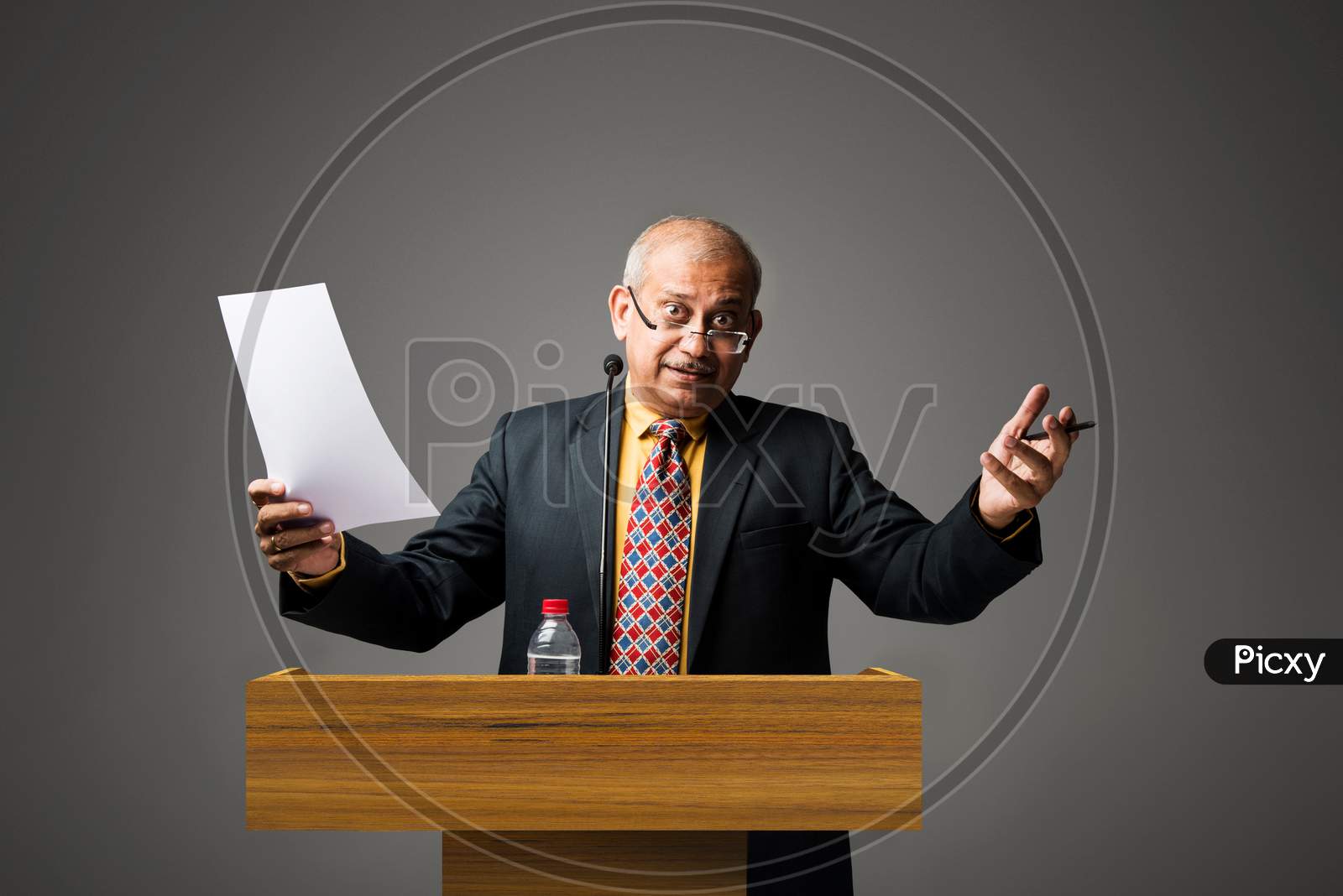 Happy Indian/asian handsome senior businessman speaking with mic at podium in office or auditorium presenting something