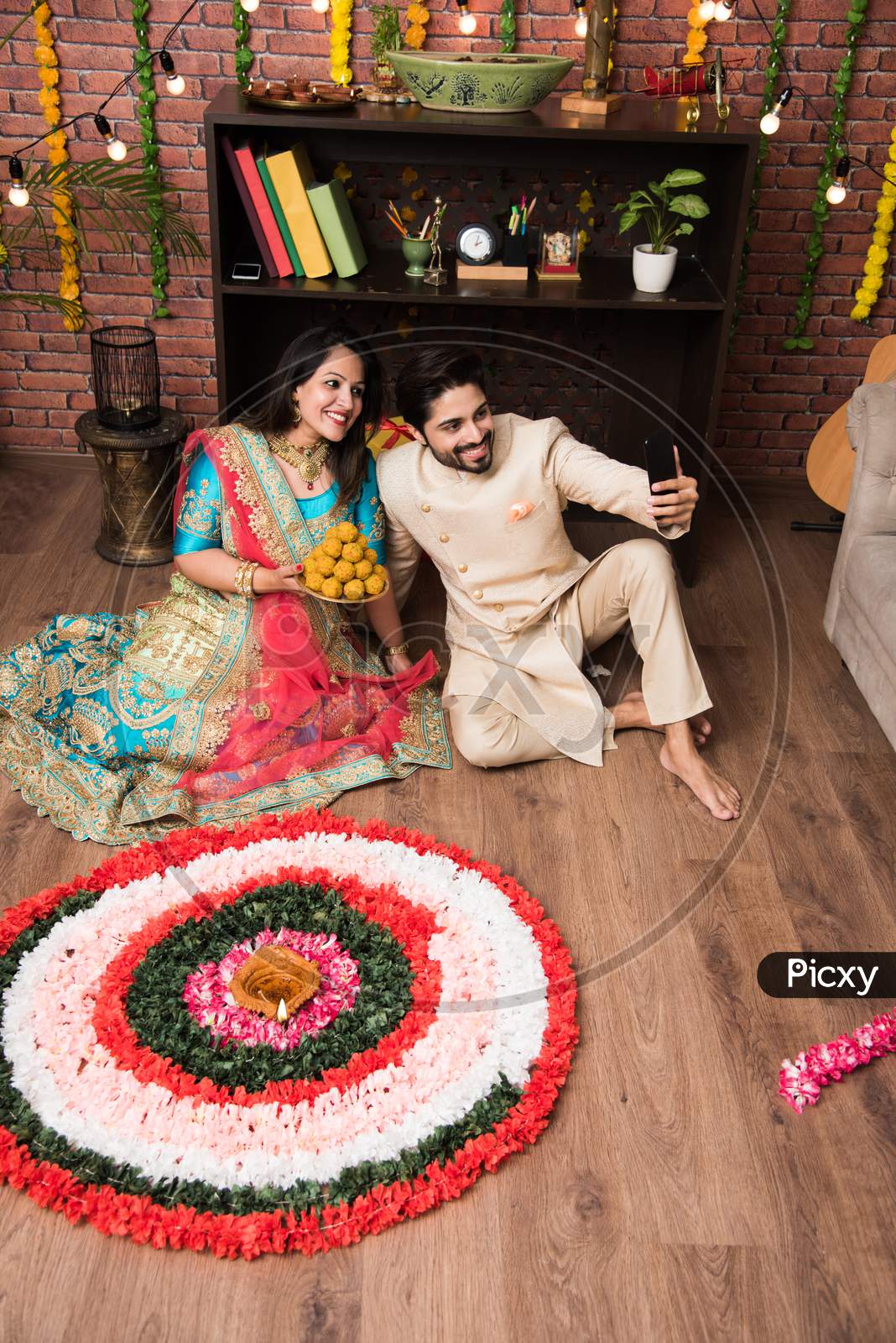 Indian couple making flower Rangoli on Diwali or Onam Festival, taking selfie or holding sweets