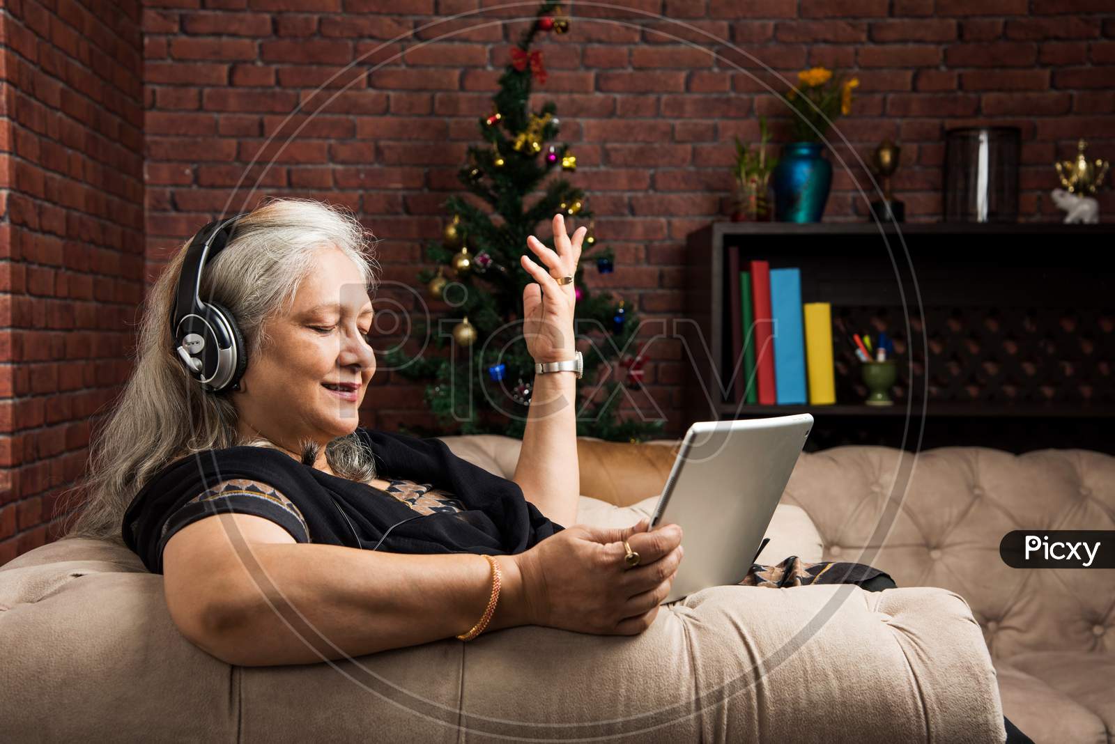 senior Indian/asian women listening to music using headphones while holding a tablet computer and sitting on sofa or couch at ho